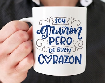 May include: White ceramic mug with a blue and gray design. The mug features the text "Soy gruñón pero de buen corazón" which translates to "I'm grumpy but good-hearted".