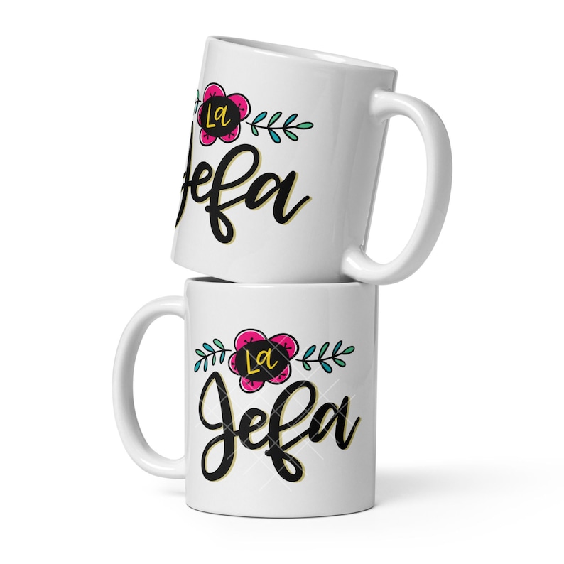 May include: Two white ceramic mugs stacked on top of each other. The mugs have a black floral design with the text "La Jefa" in gold and pink.