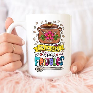 May include: White ceramic coffee mug with a colorful illustration of a pot of beans, a spoon, and the text "En la casa hay frijoles".