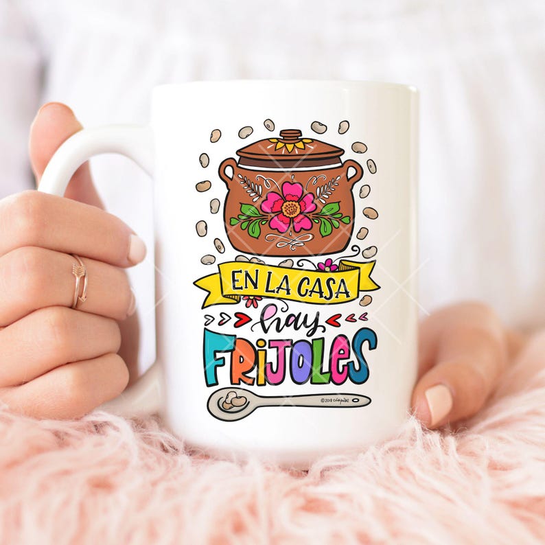 May include: White ceramic coffee mug with a colorful illustration of a pot of beans, a spoon, and the text "En la casa hay frijoles".