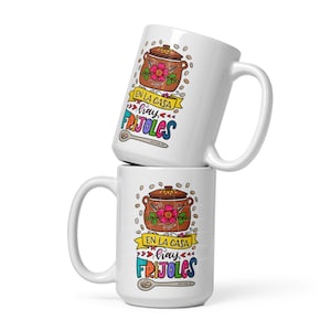 May include: Two white ceramic mugs stacked on top of each other. The mugs feature a colorful illustration of a pot of beans with the text "En la casa hay frijoles" in Spanish. The text is in a colorful, whimsical font.