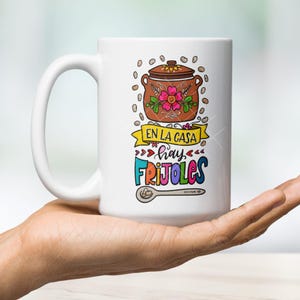 May include: White ceramic mug with a colorful illustration of a pot of beans, a spoon, and the text "En la casa hay frijoles".