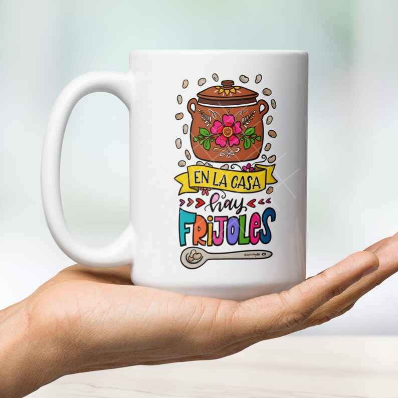 May include: White ceramic mug with a colorful illustration of a pot of beans, a spoon, and the text "En la casa hay frijoles".