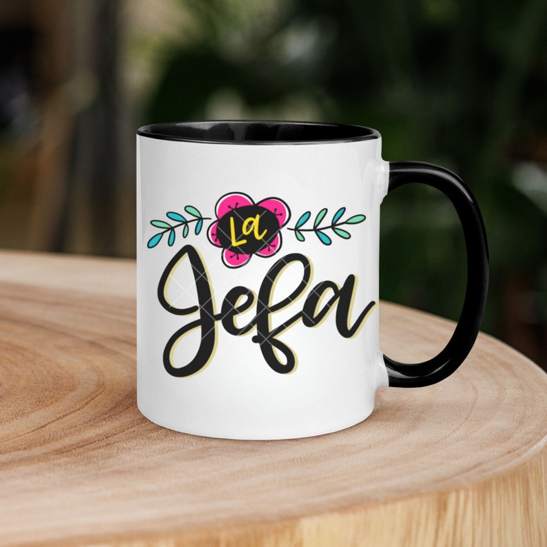 May include: White ceramic mug with a black rim and handle. The mug has a floral design with a pink flower and green leaves. The text "La Jefa" is written in black cursive lettering.