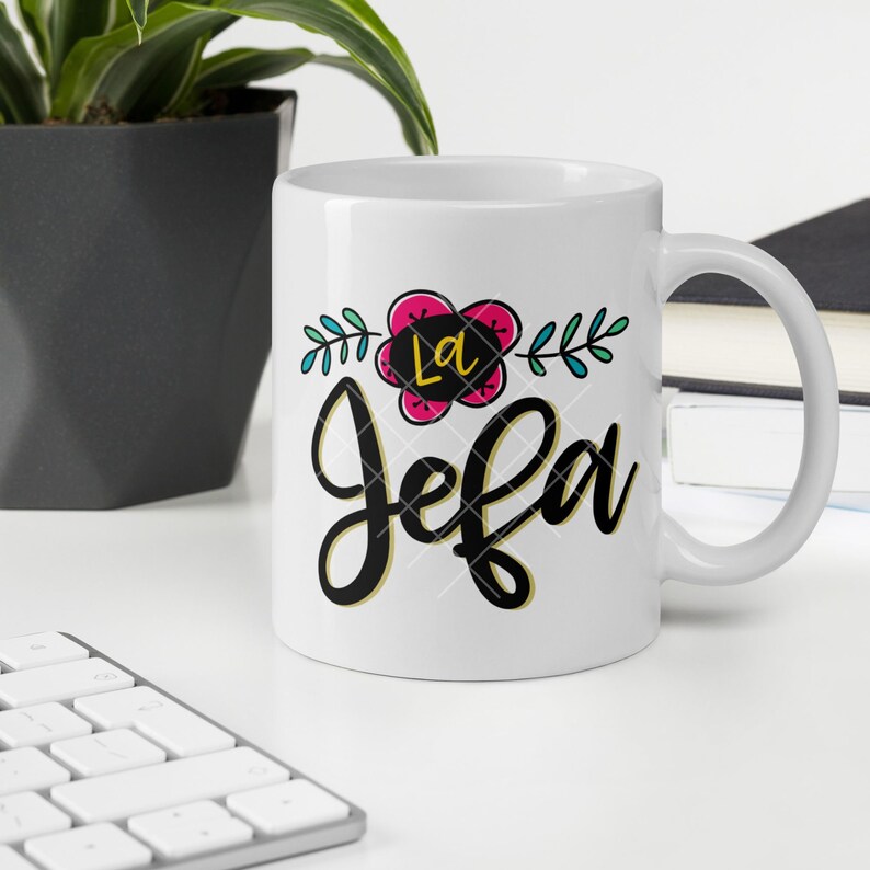 May include: White ceramic mug with a black and gold design that says "La Jefa" with a pink flower and green leaves.