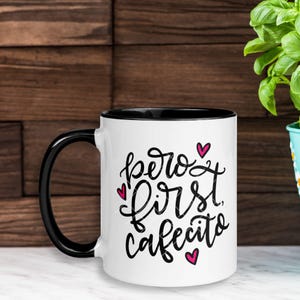 May include: White ceramic mug with a black rim and handle. The mug has a black graphic that says "pero first cafecito" with pink hearts.