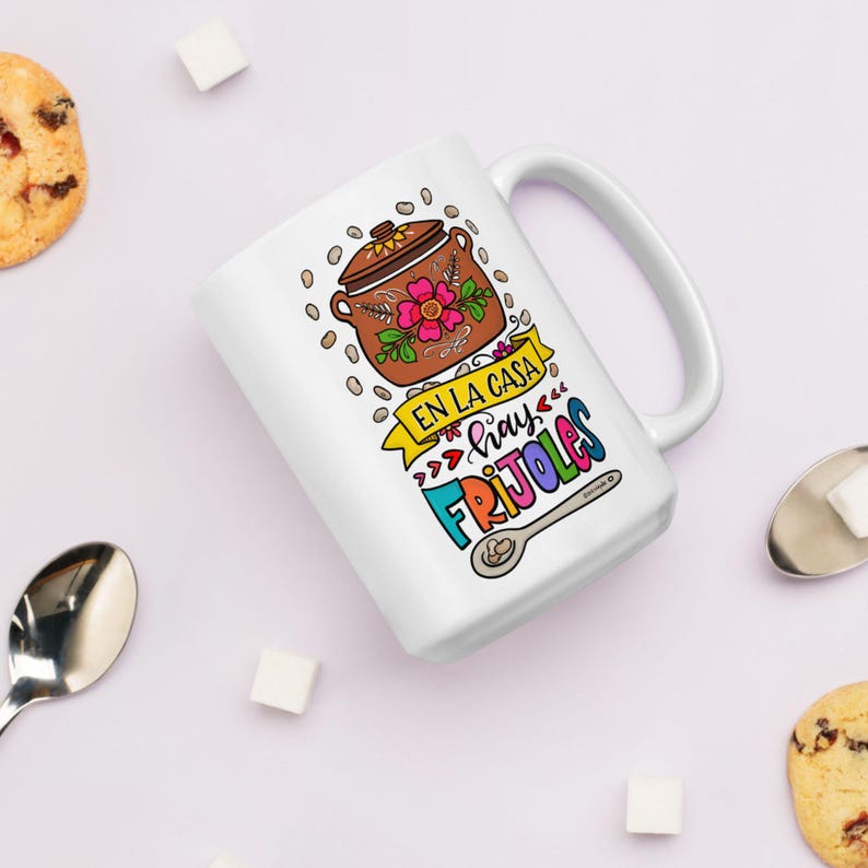 May include: White ceramic mug with a colorful illustration of a pot of beans, a spoon, and the text "En la casa hay frijoles".