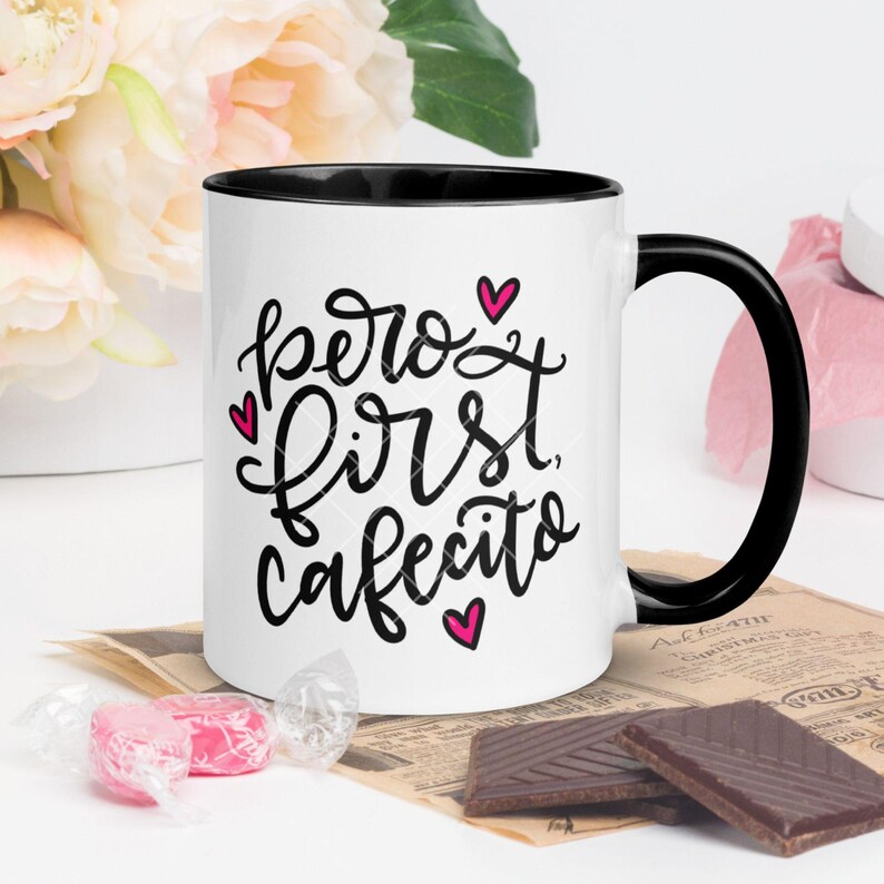 May include: White ceramic coffee mug with a black rim and handle. The mug has black lettering that says "pero first, cafecito" with pink hearts.