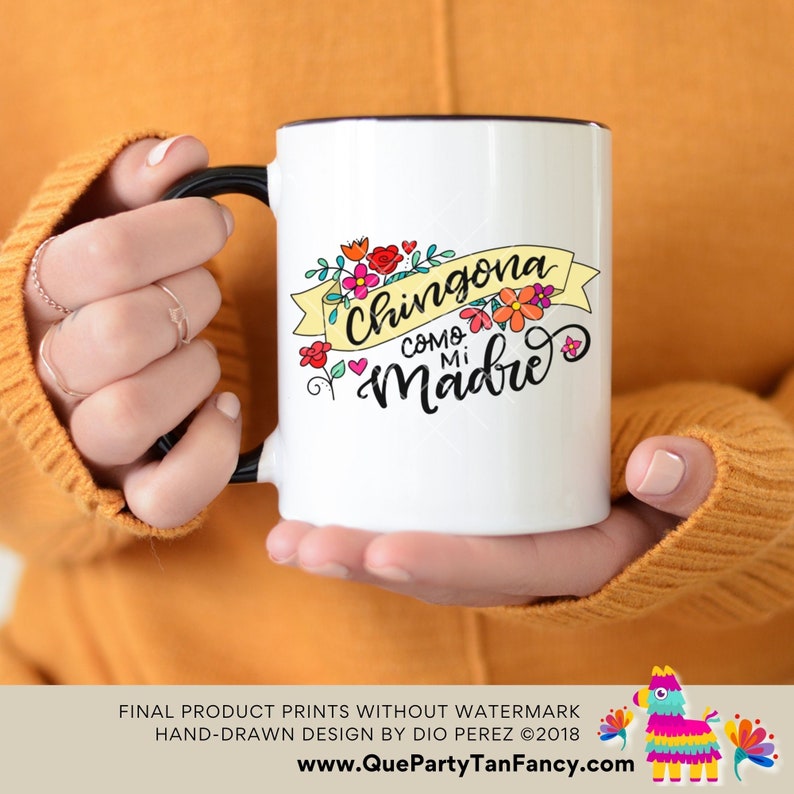 May include: White ceramic coffee mug with a black handle and black rim. The mug features a colorful floral design and the text "Chingona como mi Madre" in black lettering.