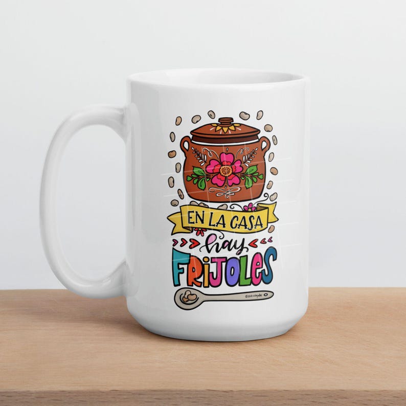 May include: White ceramic mug with a colorful illustration of a pot of beans and the text "En la casa hay frijoles".