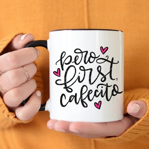 May include: White ceramic mug with a black handle and black rim. The mug has a pink and black design that says "pero first, cafecito" with pink hearts.