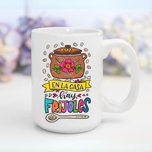 May include: White ceramic mug with a colorful illustration of a pot of beans, a spoon, and the text "En la casa hay frijoles".