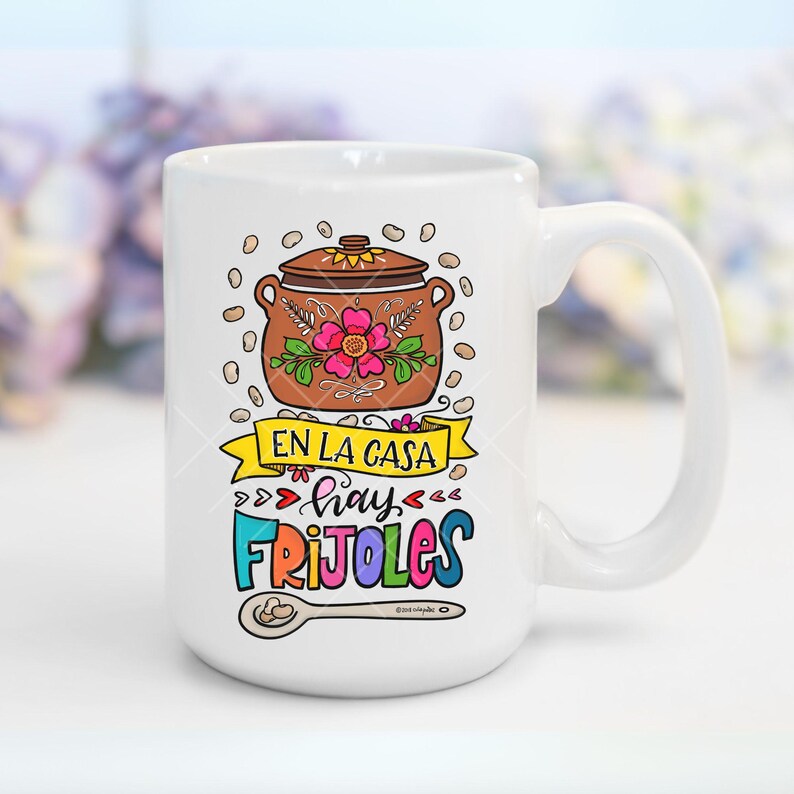 May include: White ceramic mug with a colorful illustration of a pot of beans, a spoon, and the text "En la casa hay frijoles".