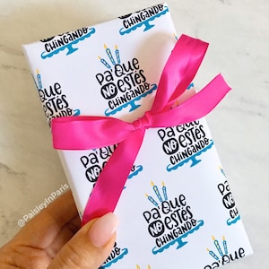 Könnte beinhalten: Ein verpacktes Geschenk mit weißem Papier mit einem sich wiederholenden Muster eines Kuchens mit Kerzen und dem Text "Pa' Que No Estés Chingando" in Schwarz und Blau. Ein leuchtend rosa Band ist um das Geschenk gebunden.