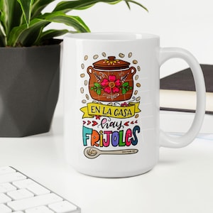May include: White ceramic mug with a colorful illustration of a pot of beans, a spoon, and the text "En la casa hay frijoles".