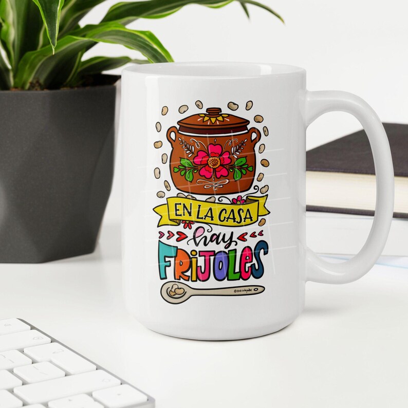 May include: White ceramic mug with a colorful illustration of a pot of beans, a spoon, and the text "En la casa hay frijoles".