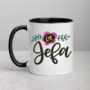 May include: White ceramic mug with a black rim and handle. The mug features a black and yellow graphic that says "La Jefa" with a pink flower and green leaves.