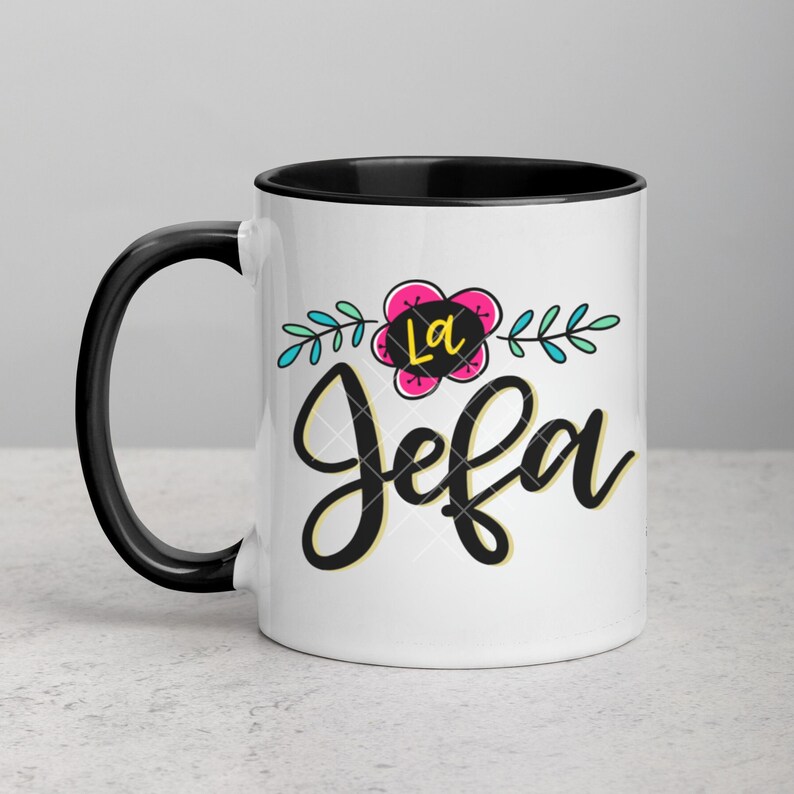May include: White ceramic mug with a black rim and handle. The mug features a black and yellow graphic that says "La Jefa" with a pink flower and green leaves.