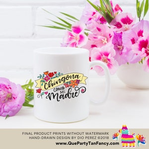 May include: White ceramic mug with a colorful graphic design that says "Chingona Como Mi Madre" with flowers and a yellow ribbon. The mug is sitting on a white surface with pink flowers in the background.