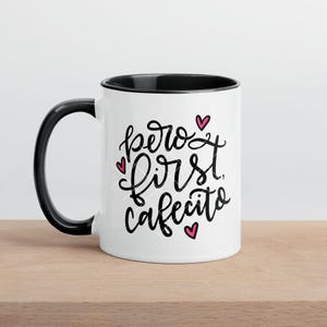 May include: White ceramic coffee mug with a black rim and handle. The mug has black text that reads "pero first cafecito" with pink hearts.