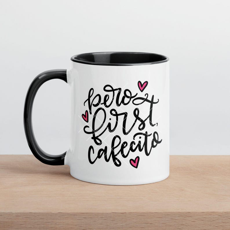 May include: White ceramic coffee mug with a black rim and handle. The mug has black text that reads "pero first cafecito" with pink hearts.