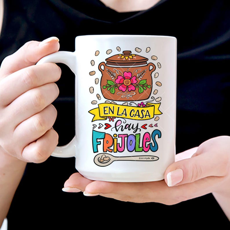 May include: White ceramic mug with a colorful illustration of a pot of beans, a spoon, and the text "En la casa hay frijoles".