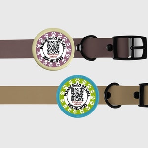 Puede incluir: Dos collares para mascotas con etiquetas circulares. Las etiquetas tienen un código QR, el texto «PLEASE SCAN OR CALL» y un número de teléfono. Una etiqueta es lavanda con huellas de patas blancas, la otra es turquesa con huellas de patas blancas. Los collares son marrones y beige.