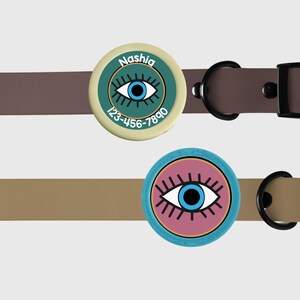 May include: Two round dog tags with a blue eye design and black eyelashes. The top tag is green and has the name "Nashia" and the phone number "123-456-7890". The bottom tag is pink.