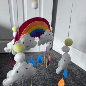 May include: A rainbow-themed baby mobile with white clouds, yellow pom-poms, and colorful raindrops hanging from a wooden hoop.