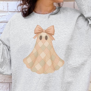 May include: Pale grey sweatshirt with a ghost design. The ghost has a bow and a peach and cream plaid pattern. The word "ASH" is printed in the bottom left corner of the image.