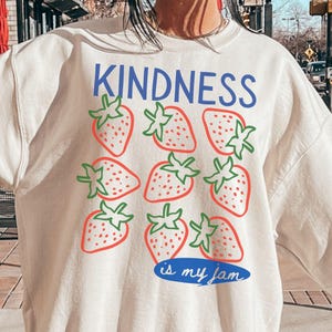Peut inclure: Sweat-shirt couleur sable avec le mot "KINDNESS" en bleu au-dessus d'un motif de fraises rouges à tiges vertes. L'expression "is my jam" est écrite en bleu sous les fraises.