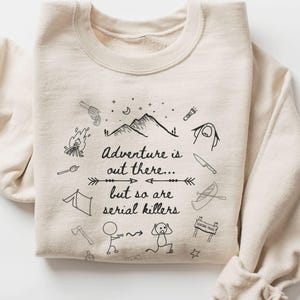 May include: Sand-colored sweatshirt with the text "Adventure is out there... but so are serial killers" and various black line-drawn illustrations of outdoor adventure elements. The illustrations include a mountain, tent, campfire, and a canoe.