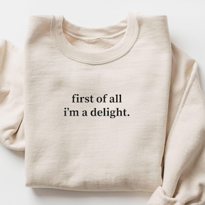 May include: A sand-coloured sweatshirt with the text "first of all i'm a delight." The long-sleeved top is folded neatly. The word "SAND" is in a rectangular box in the lower left corner.