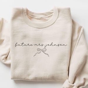 May include: A sand-coloured sweatshirt featuring the script "future mrs. johnson" and a small bow illustration. The sleeves are rolled up. The design is simple and elegant, suitable for bridal wear, with a focus on a minimalist aesthetic.