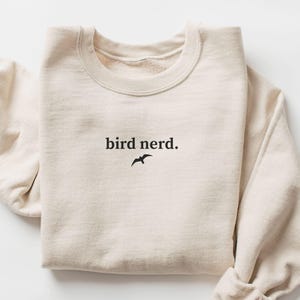 May include: A sand-coloured sweatshirt featuring the words "bird nerd." in black, with a small bird silhouette. The long-sleeved top is folded, displaying a casual, comfortable style. The word "SAND" is in a box in the lower left corner.