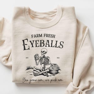 May include: Sand-coloured sweatshirt featuring a black graphic. The graphic reads "FARM FRESH EYEBALLS" above a skeleton holding a jar and sitting next to a basket of eyeballs. The text "You grow em, we pick em" is below the skeleton.