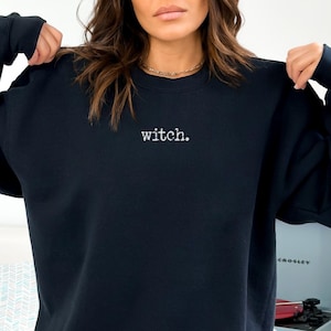May include: Black sweatshirt with the word "witch." embroidered in white. The sweatshirt is a crew neck style. The word "BLACK" is in a black rectangle in the lower left corner.