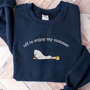 May include: Navy blue sweatshirt featuring the text "off to enjoy my summer". An illustration of a goose with a yellow mug is also present. The sweatshirt has a crew neck and long sleeves.