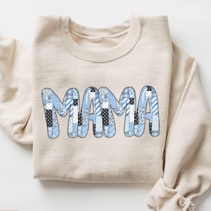 Peut inclure: Un sweat-shirt couleur sable avec le mot "MAMA" en patchwork de motifs bleus et blancs. Les motifs comprennent des rayures, des pois et des carreaux. Le sweat-shirt est plié.