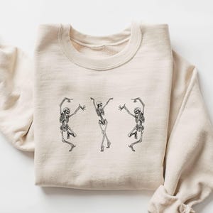 May include: A sand-coloured sweatshirt with a black graphic of three dancing skeletons. The skeletons are in a playful pose, with arms and legs outstretched. The sweatshirt is folded neatly, showcasing the design.