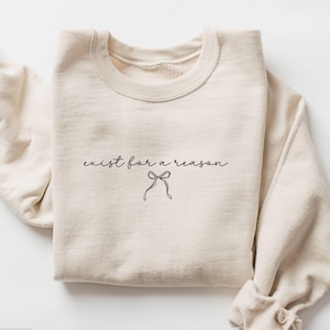 May include: Sand-colored sweatshirt with the phrase "exist for a reason" in a delicate script, accompanied by a small bow illustration. The sweatshirt is folded neatly, showcasing its soft texture and casual style.