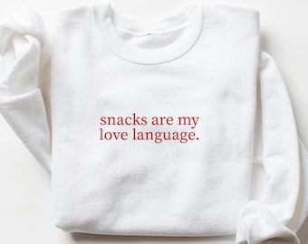 May include: White sweatshirt with the phrase "snacks are my love language" in red lettering. The long-sleeved top is made of a soft material and is a casual style. The word "WHITE" is printed at the bottom of the image.