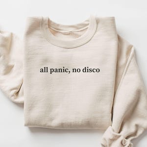 May include: A sand-colored sweatshirt with the phrase "all panic, no disco" printed in black. The long-sleeved top is folded neatly, with the sleeves rolled up. The word "SAND" is printed in a rectangular box in the bottom left corner.