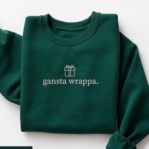 May include: A forest green sweatshirt featuring the white text "gansta wrappa." and a gift box graphic. The garment is neatly folded, displaying the design. The words "FOREST GREEN" are displayed at the bottom of the image.