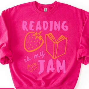 May include: A vibrant pink sweatshirt featuring the phrase "READING is my JAM" in pink and orange. The design includes a strawberry and an open book. The brand name "HELICONA" is in a white box in the lower left corner.
