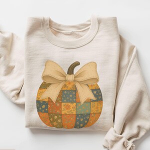 May include: A sand-coloured sweatshirt with a patchwork pumpkin design and a large bow. The pumpkin is made of squares in orange, green, and yellow with floral patterns. The sweatshirt is folded, with the word "SAND" at the bottom.