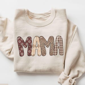 May include: A sand-coloured sweatshirt displaying the word "MAMA" in a patchwork design. The letters feature patterns such as snowflakes, argyle, leopard print, and gingham. The sweatshirt is neatly folded.
