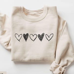 May include: A sand-colored sweatshirt with a crew neck and long sleeves. The front features five black sketched hearts in a row. The sweatshirt is folded, with the sleeves cuffed at the wrists. The word "SAND" is in a rectangular box in the lower left corner.