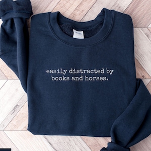 May include: A navy blue sweatshirt with the white text "easily distracted by books and horses". The garment is folded on a light wood-effect surface. The word "NAVY" is printed in the lower left corner.