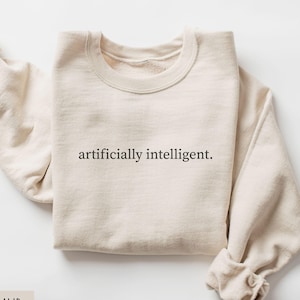May include: A sand-coloured crewneck sweatshirt featuring the text "artificially intelligent." printed in a black serif font across the centre of the chest. The garment has long sleeves with ribbed cuffs and a ribbed neckline, presented in a flat lay.